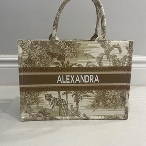 May include: A beige tote bag with a brown stripe featuring the name "ALEXANDRA" in white. The bag has a jungle-themed pattern with a tiger and palm trees. The handles are beige with a matching pattern.