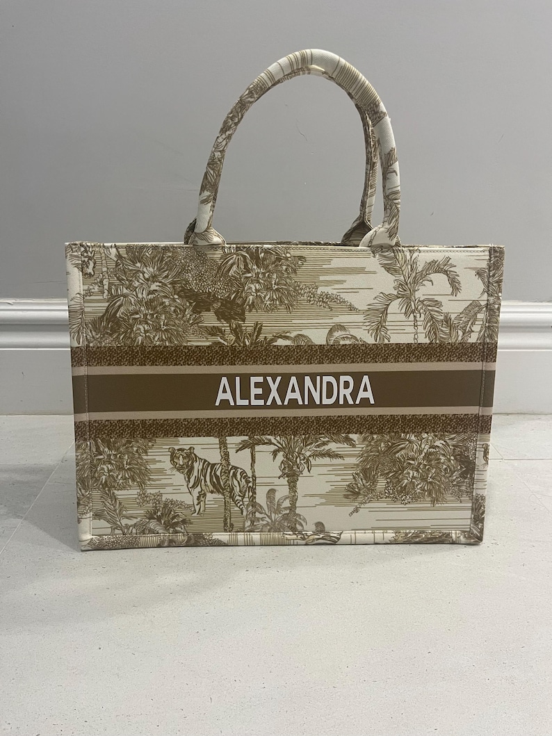 May include: A beige tote bag with a brown stripe featuring the name "ALEXANDRA" in white. The bag has a jungle-themed pattern with a tiger and palm trees. The handles are beige with a matching pattern.