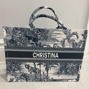 May include: A tote bag with a black and white jungle print featuring a tiger and palm trees. The bag has two handles and a black stripe across the front with the name "CHRISTINA" in white letters.
