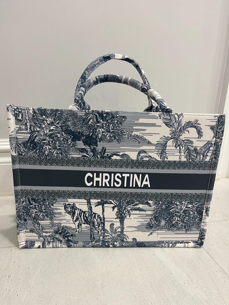 May include: A tote bag with a black and white jungle print featuring a tiger and palm trees. The bag has two handles and a black stripe across the front with the name "CHRISTINA" in white letters.