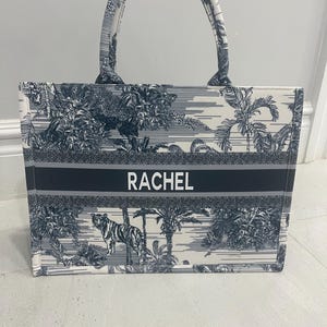 May include: A rectangular tote bag with a white and navy blue tropical print featuring a tiger. The bag has a navy blue stripe with the name "RACHEL" in white. The handles and trim are navy blue.