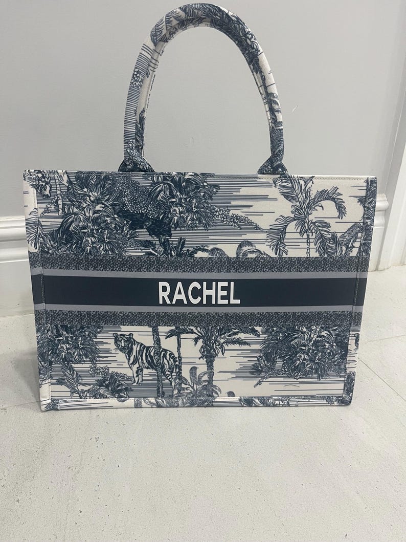 May include: A rectangular tote bag with a white and navy blue tropical print featuring a tiger. The bag has a navy blue stripe with the name "RACHEL" in white. The handles and trim are navy blue.