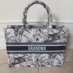 May include: A tote bag with a black and white jungle print featuring leopards and foliage. The bag has a black and white striped band with the word "GRANDMA" in white. It has a zipper closure and two handles.
