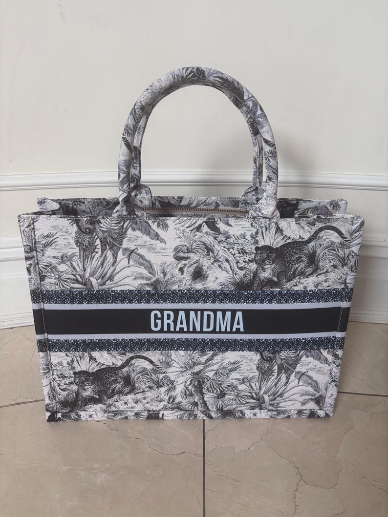 May include: A tote bag with a black and white jungle print featuring leopards and foliage. The bag has a black and white striped band with the word "GRANDMA" in white. It has a zipper closure and two handles.