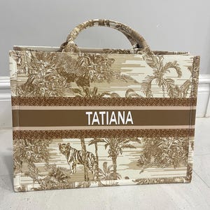 May include: A beige tote bag with a brown jungle-themed pattern featuring a tiger, palm trees, and other foliage. The bag has a brown stripe with the name "TATIANA" in white letters. The handles and trim are also brown.