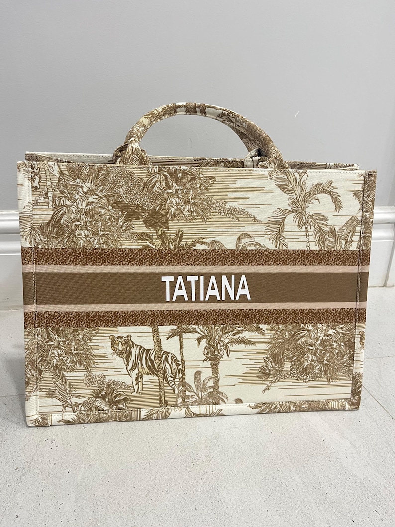May include: A beige tote bag with a brown jungle-themed pattern featuring a tiger, palm trees, and other foliage. The bag has a brown stripe with the name "TATIANA" in white letters. The handles and trim are also brown.