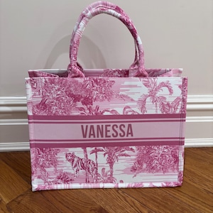 May include: Pink and white tote bag with a tropical print featuring a tiger. The bag has a striped handle and the name "VANESSA" printed on the front. The bag is made of a sturdy material and is suitable for carrying various items.