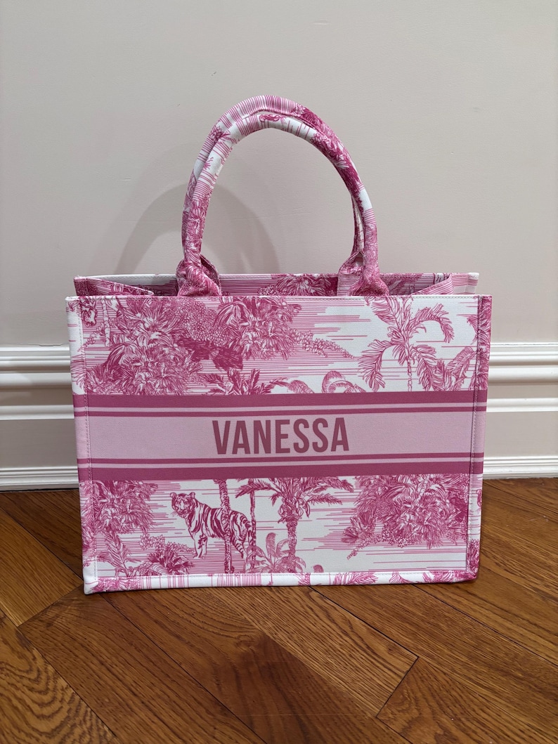 May include: Pink and white tote bag with a tropical print featuring a tiger. The bag has a striped handle and the name "VANESSA" printed on the front. The bag is made of a sturdy material and is suitable for carrying various items.