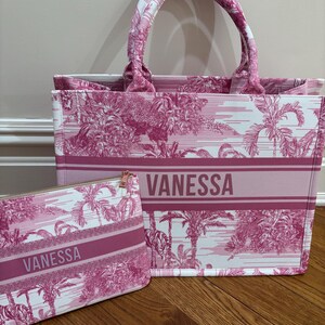 May include: A pink and white tote bag and matching zippered pouch. The bags feature a repeating tropical pattern and the name "VANESSA" in a bold font. The tote bag has two handles and a rectangular shape.