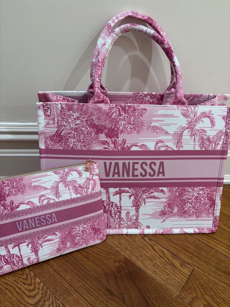 May include: A pink and white tote bag and matching zippered pouch. The bags feature a repeating tropical pattern and the name "VANESSA" in a bold font. The tote bag has two handles and a rectangular shape.