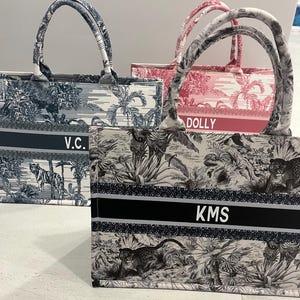 May include: Three tote bags with matching handles. One bag is black and white with a jungle scene and the initials "KMS". Another is navy blue and white with a landscape scene and the initials "V.C.". The third is pink and white with a landscape scene and the name "DOLLY".