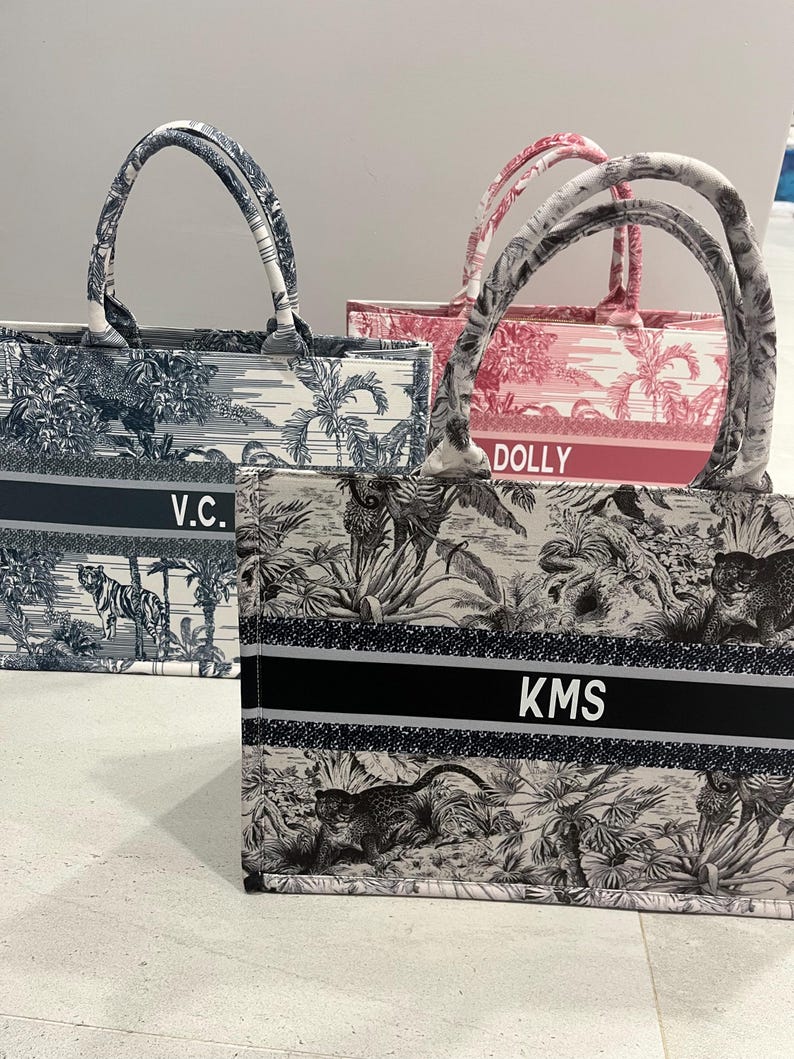 May include: Three tote bags with matching handles. One bag is black and white with a jungle scene and the initials "KMS". Another is navy blue and white with a landscape scene and the initials "V.C.". The third is pink and white with a landscape scene and the name "DOLLY".