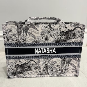 May include: A tote bag with a black and white jungle print featuring leopards and foliage. The bag has a black stripe with the name "NATASHA" in white letters. It has two handles and a rectangular shape.