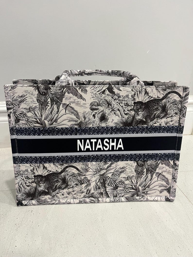 May include: A tote bag with a black and white jungle print featuring leopards and foliage. The bag has a black stripe with the name "NATASHA" in white letters. It has two handles and a rectangular shape.