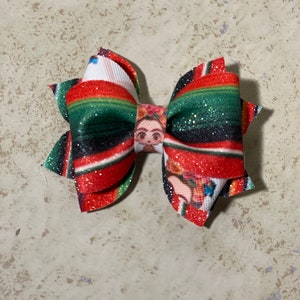 May include: A red, green, and black striped hair bow with a glitter finish. The center of the bow features a printed image of Frida Kahlo.