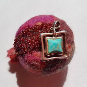 Può includere: Un pendente in argento con una pietra turchese quadrata incastonata in una doppia cornice quadrata. Il pendente poggia su un oggetto rotondo rosa scuro e strutturato con una sezione di perline di perle rosse.