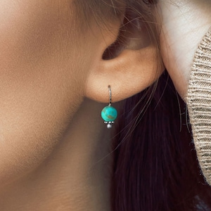 May include: A close-up of a woman's ear with a turquoise gemstone earring. The earring is silver and has a small bead at the bottom.
