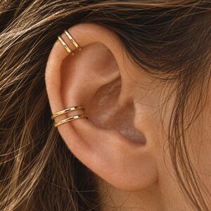May include: Two gold-toned ear cuffs worn on the upper cartilage and the conch area of the ear. Each cuff features a double-band design with a smooth, polished finish. The jewelry is positioned on the ear, showcasing the metallic gold color.