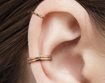 May include: Two gold ear cuffs worn on the upper part of the ear. The ear cuffs are thin and have a hammered texture.