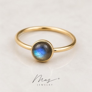 Könnte beinhalten: Ein goldener Ring mit einem runden, irisierenden Edelstein. Der Edelstein zeigt Blau-, Grün- und Grautöne. Der Ring hat ein schmales Band und ein schlichtes Design. Der Markenname "Maz Jewelry" steht darunter in Schreibschrift.