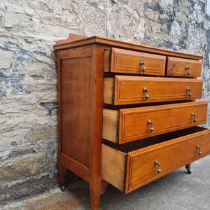 May include: A wooden dresser with five drawers, some open, showcasing a warm, honey-toned finish. The drawers have brass pulls and decorative trim. The dresser is set against a textured stone wall and has small wheels on the bottom.