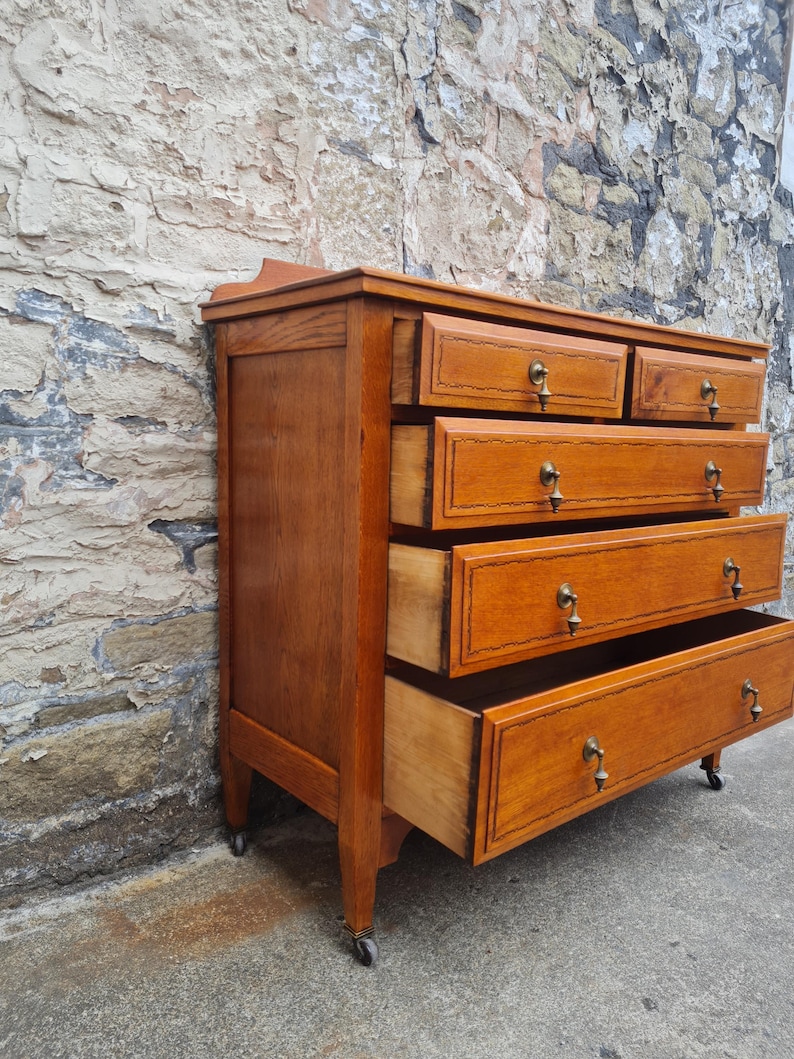 May include: A wooden dresser with five drawers, some open, showcasing a warm, honey-toned finish. The drawers have brass pulls and decorative trim. The dresser is set against a textured stone wall and has small wheels on the bottom.