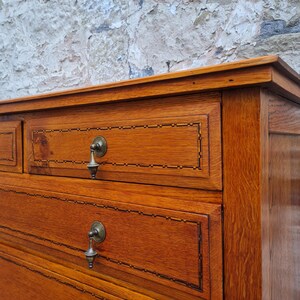 May include: Close-up of a wooden dresser with multiple drawers. The dresser is made of a warm-toned wood with visible grain. Each drawer has a decorative border and a metal pull. The background is a textured, light-colored wall.