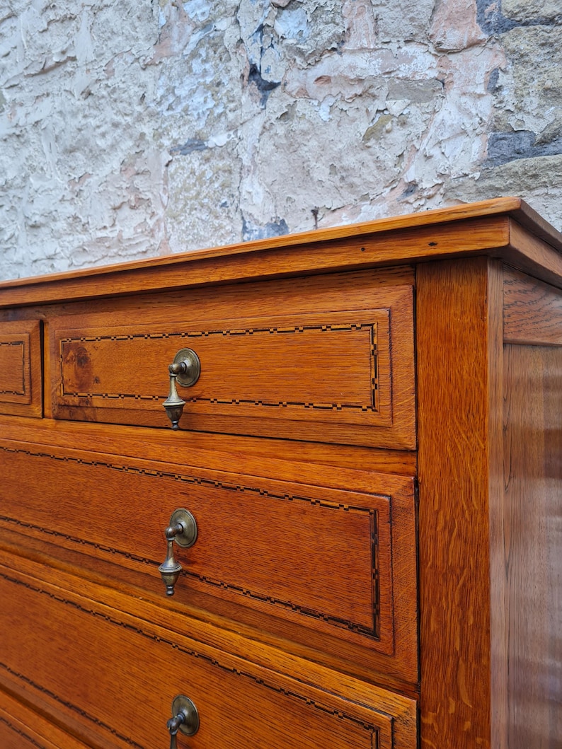 May include: Close-up of a wooden dresser with multiple drawers. The dresser is made of a warm-toned wood with visible grain. Each drawer has a decorative border and a metal pull. The background is a textured, light-colored wall.