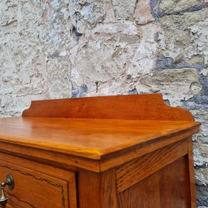 May include: Close-up of a wooden dresser with a warm, reddish-brown finish. The dresser features a decorative top, drawers with brass hardware, and detailed inlaid trim. The wood grain is visible, showcasing the craftsmanship.