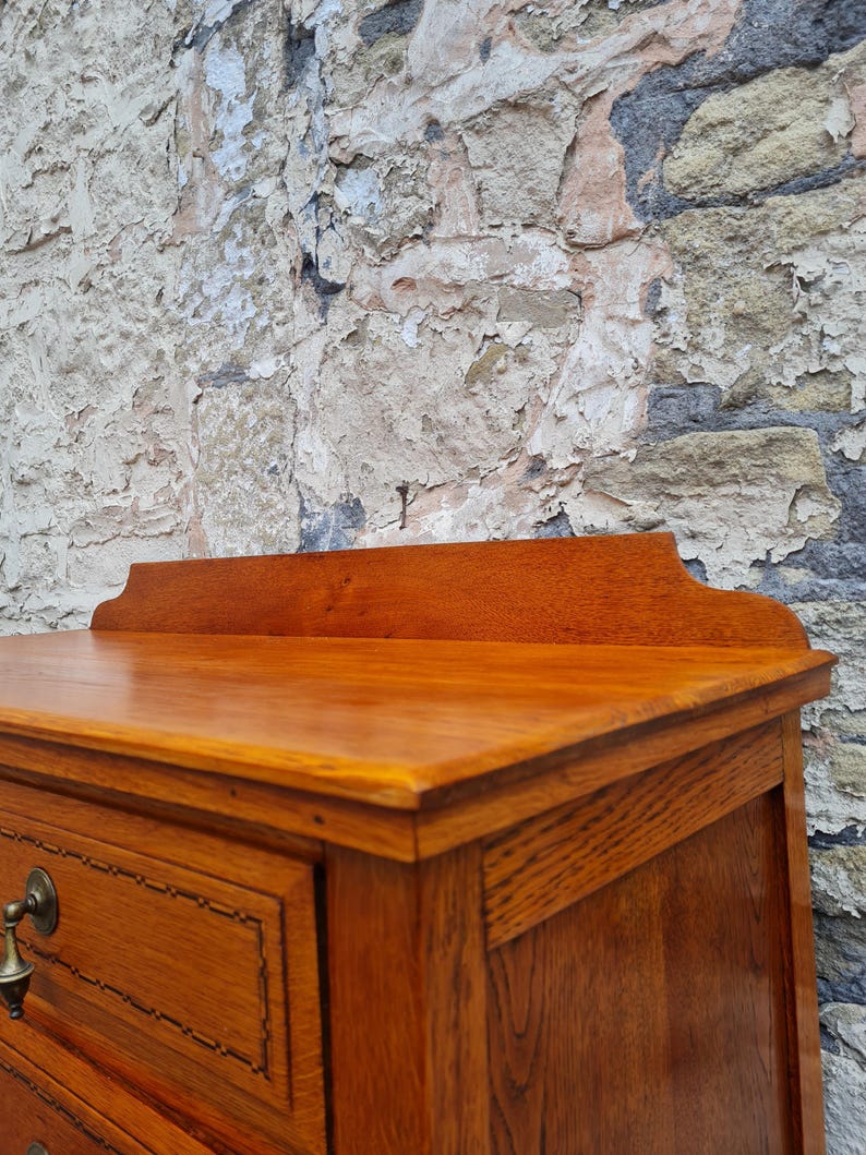 May include: Close-up of a wooden dresser with a warm, reddish-brown finish. The dresser features a decorative top, drawers with brass hardware, and detailed inlaid trim. The wood grain is visible, showcasing the craftsmanship.