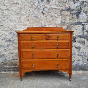 May include: A wooden chest of drawers with five drawers and brass hardware. The chest is a warm, medium brown color and has a decorative top edge. The drawers have small, round brass knobs. The chest sits on small wheels.