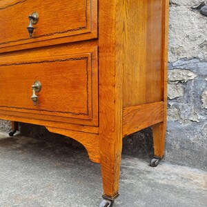 May include: A close-up of a wooden dresser with three drawers. The dresser is a warm, reddish-brown color with brass hardware and decorative trim around the drawers. The dresser sits on small wheels.