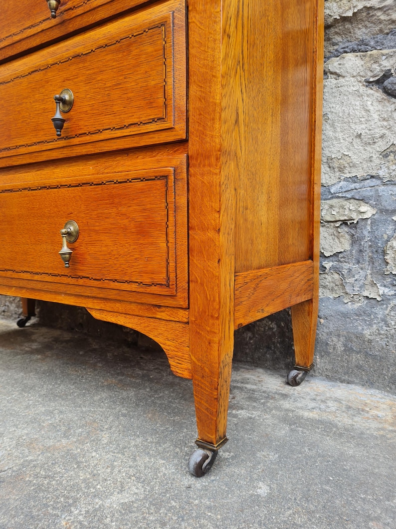 May include: A close-up of a wooden dresser with three drawers. The dresser is a warm, reddish-brown color with brass hardware and decorative trim around the drawers. The dresser sits on small wheels.
