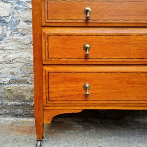 May include: A close-up of a wooden dresser with four drawers. The dresser is made of light brown wood with brass handles and decorative trim. The bottom of the dresser has a curved design, and the legs have small wheels.