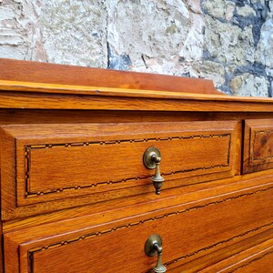 May include: Close-up of a wooden dresser with two drawers. The dresser is a warm, reddish-brown color with brass drawer pulls. The drawers have a decorative inlaid border. The background is a textured stone wall.