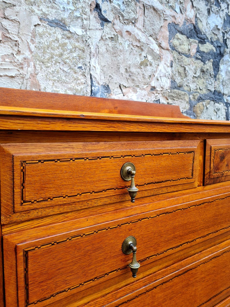 May include: Close-up of a wooden dresser with two drawers. The dresser is a warm, reddish-brown color with brass drawer pulls. The drawers have a decorative inlaid border. The background is a textured stone wall.