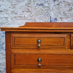 May include: A close-up of a wooden dresser with three drawers. The dresser is made of light brown wood with a decorative border around each drawer. Each drawer has a brass pull. The background is a textured brick wall.