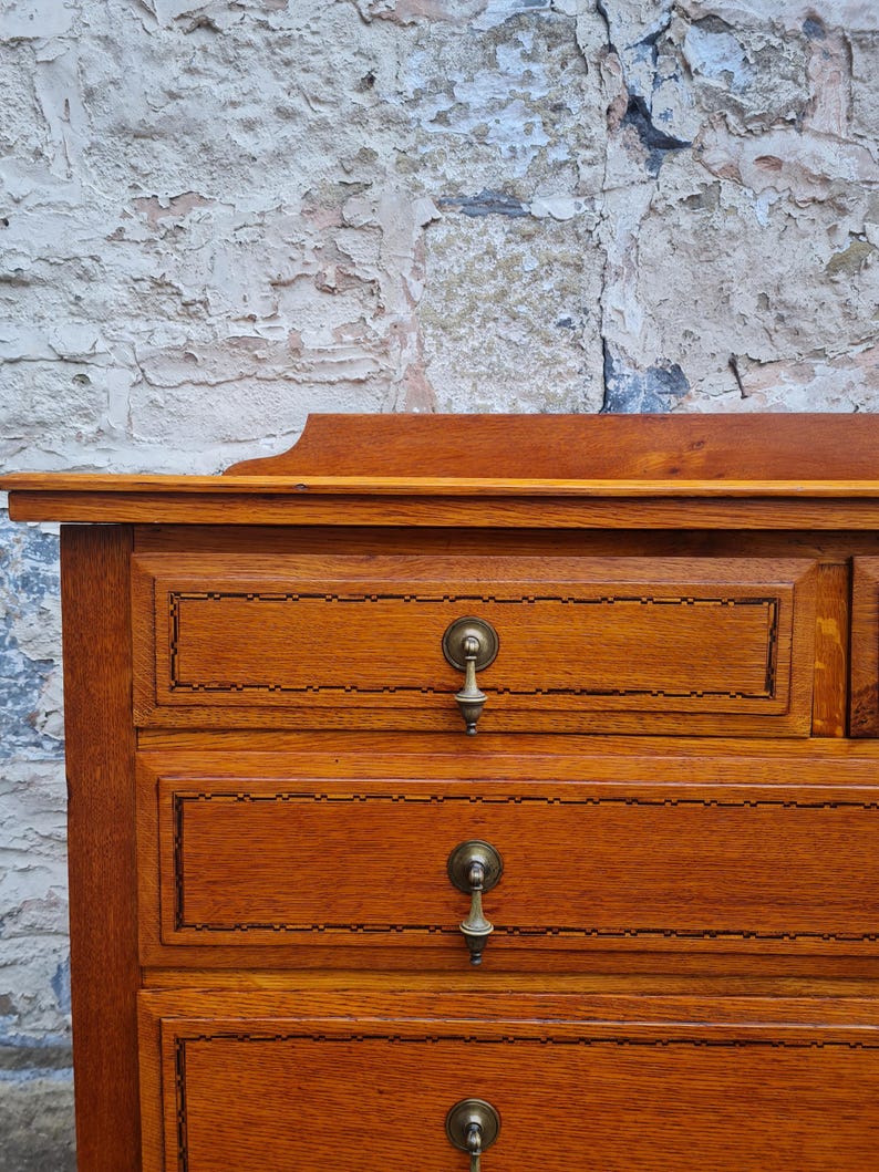 May include: A close-up of a wooden dresser with three drawers. The dresser is made of light brown wood with a decorative border around each drawer. Each drawer has a brass pull. The background is a textured brick wall.