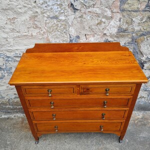 May include: A wooden dresser with a warm, amber finish. It features five drawers with brass knobs and sits on small wheels. The top has a slightly curved edge. The dresser is against a textured, weathered stone wall.