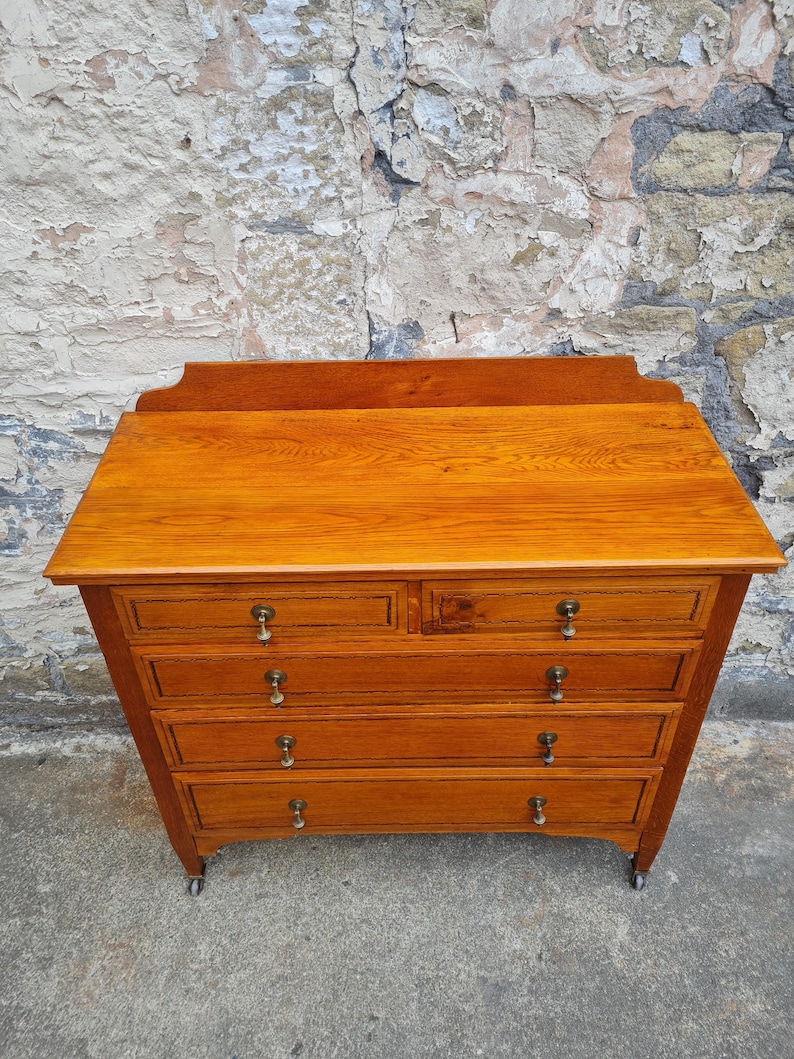 May include: A wooden dresser with a warm, amber finish. It features five drawers with brass knobs and sits on small wheels. The top has a slightly curved edge. The dresser is against a textured, weathered stone wall.