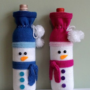 以下が含まれることがあります： 2つの編み物雪だるまワインボトルカバー。1つは青い帽子、スカーフ、ボタン、オレンジ色のニンジン鼻が付いています。もう1つはピンクの帽子、スカーフ、ボタン、オレンジ色のニンジン鼻が付いています。どちらも白いポンポンが付いています。