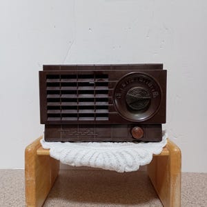 Könnte beinhalten: Ein dunkelbraunes, rechteckiges Radio mit einem horizontalen Lamellen-Lautsprechergitter auf der linken Seite und einem runden Abstimmknopf auf der rechten Seite. Das Radio hat einen kleinen runden Drehknopf unten rechts und den Markennamen Motorola.