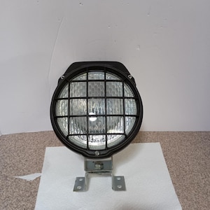 May include: A black round spotlight with a protective grid, mounted on a metal bracket. The light has a clear lens and is designed for vehicle or equipment use.