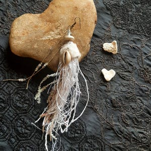 Può includere: Orecchino fatto a mano con conchiglia e frange bianche, presentato su una pietra marrone chiaro. L'orecchino è appeso a un gancio d'argento. Sono presenti anche due piccole pietre a forma di cuore. Lo sfondo è un tessuto scuro e strutturato.