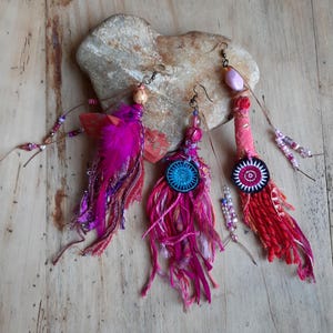 May include: Three bohemian-style earrings. Each earring is crafted with colourful fabric, beads, and feathers. The earrings feature shades of pink, red, and purple, with decorative buttons and dangling accents. The earrings are displayed on a wooden surface.