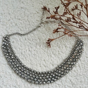 May include: A silver choker necklace with multiple rows of clear rhinestones. The necklace is on a white fluffy surface.
