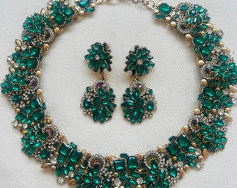 Puede incluir: Un collar de declaración dorado y verde esmeralda y pendientes a juego. El collar presenta un diseño grande y ornamentado con una variedad de piedras verdes esmeralda y detalles dorados. Los pendientes también son grandes y presentan un diseño similar.