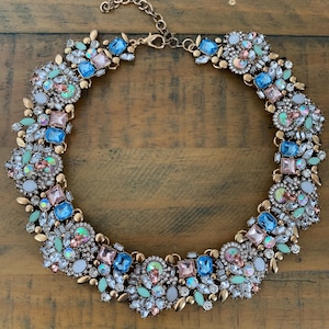 May include: A statement necklace with a gold-tone chain, adorned with blue, pink, and green gemstones. The gemstones are arranged in a floral pattern. The necklace is designed to be a bold accessory.