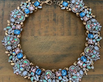 Puede incluir: Collar llamativo con piedras preciosas azules, rosas y verdes. La cadena y el cierre son dorados. Las piedras están dispuestas en un diseño floral, creando una joya audaz.