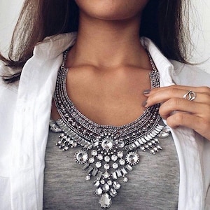 May include: Silver statement necklace with clear crystal embellishments. The necklace has a bib design with multiple layers of sparkling stones and detailed metalwork. It is worn over a grey top and a white shirt.